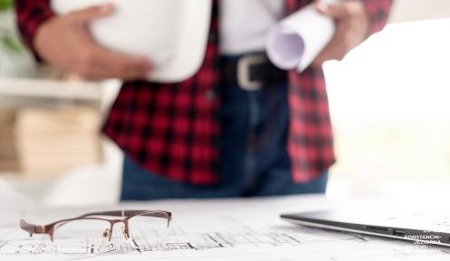 Mężczyzna z kaskiem w jednej i projektem w drugiej ręce stoi przy stole, na którym leży inny projekt i okulary