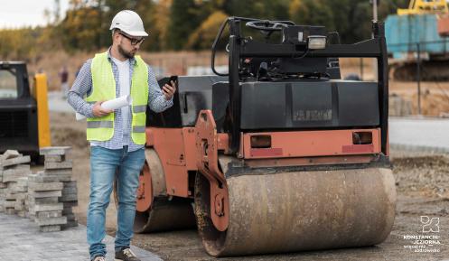 Robotnik w kamizelce odblaskowej i kasku stoi obok walca drogowego na placu budowy drogi, trzymając dokumentację i telefon.