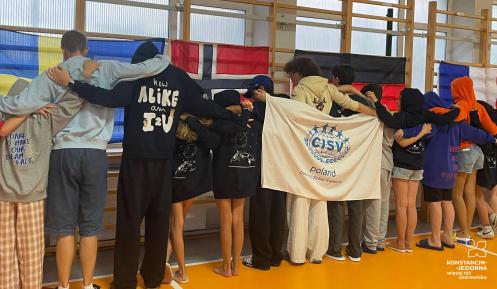 Grupa młodzieży stoi tyłem w szpalerze, obejmując się ramionami w hali sportowej. Przed nimi powieszona jest biała flaga z logo organizacji oraz napisem „CISV poland building global friendship”. W tle na szkolnych drabinkach wiszą flagi państwowe, m.in. Szwecji, Norwegii, Niemiec i Francji.