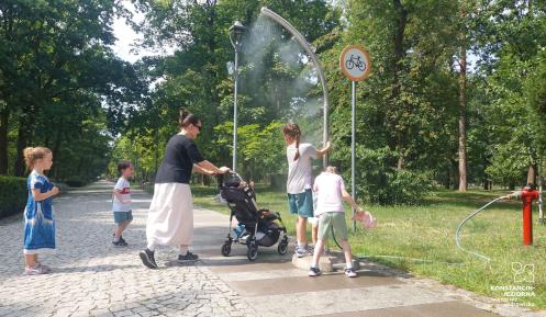 Na parkowej alei kobieta z dziećmi korzysta z bramki zraszającej, która rozpylając wodną mgiełkę przynosi ochłodę podczas upalnego dnia.