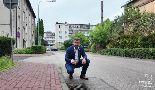 Burmistrz kuca przy wpuście kanalizacji deszczowej na osiedlowej ulicy, wskazując element infrastruktury przeznaczonej do modernizacji, a w tle widoczne są budynki mieszkalne i zieleń.