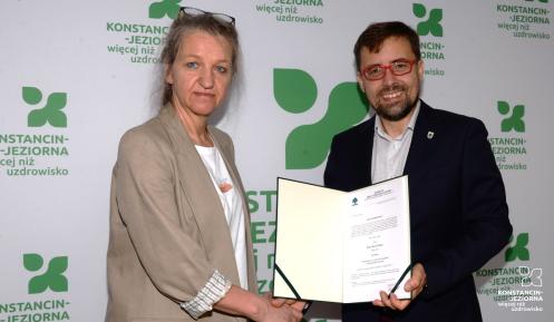Na zdjęciu kobieta (dyrektor Ewa Winek) i mężczyzna (burmistrz Michał Wiśniewski) stoją przed ścianką z logo gminy Konstancin-Jeziorna. Uściskiem dłoni potwierdzają wręczenie aktu powierzenia stanowiska dyrektora. Mężczyzna trzyma otwartą teczkę z widocznym dokumentem i uśmiecha się do obiektywu.