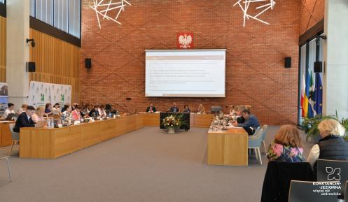 Grupa osób siedzi za stołami na sali posiedzeń