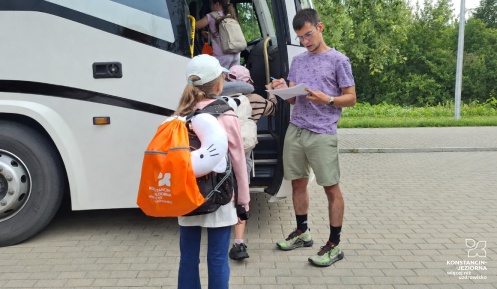 Dzieci z plecakami wsiadają do autokaru przed wyjazdem na wakacyjny wypoczynek w Zakopanem. Opiekun sprawdza listę uczestników.