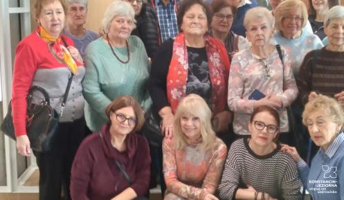 Grupa kilkunastu seniorek i kilku seniorów pozuje do wspólnego zdjęcia we wnętrzu budynku. Część osób stoi, a cztery kobiety siedzą lub klęczą z przodu.