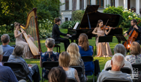 Plenerowy koncert muzyki klasycznej w ogrodzie przed zabytkową willą. Muzycy grają na harfie, fortepianie, skrzypcach i wiolonczeli, a publiczność słucha ich, siedząc na krzesłach ustawionych na trawniku