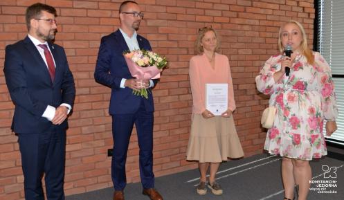 Cztery osoby stoją przed ceglaną ścianą podczas uroczystego podziękowania. Jedna z kobiet trzyma dyplom, mężczyzna bukiet kwiatów, a druga kobieta przemawia do mikrofonu