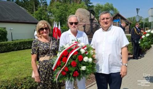 Trzyosobowa delegacja Rady Seniorów Gminy Konstancin-Jeziorna – Alfreda Konopka, Wojciech Guszkowski i Grzegorz Rutkowski – stoi z wieńcem biało-czerwonych goździków przed pomnikiem pamięci z symbolem Polski Walczącej podczas obchodów rocznicy Powstania Warszawskiego.