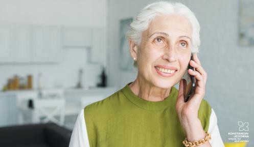 Uśmiechnięta seniorka o siwych włosach rozmawia przez telefon komórkowy. Kobieta przebywa w jasnym, domowym wnętrzu