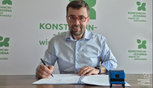 Burmistrz siedzi przy biurku i podpisuje dokumenty, a na stole obok znajdują się pieczątka oraz dokumentacja.