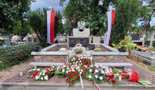 Pomnik wojenny na cmentarzu udekorowany biało-czerwonymi wieńcami i wiązankami kwiatów, fluktuujący dwoma polskimi flagami po bokach. Na płycie widnieje godło Polski oraz napis „Chwała Bohaterom”.
