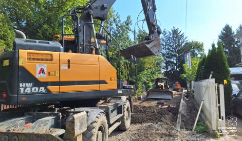 Zdjęcie przedstawia plac budowy drogi na osiedlu mieszkaniowym w słoneczny dzień. Na pierwszym planie po lewej stoi duża żółto-czarna koparka kołowa Hyundai HW140A z uniesionym ramieniem i łyżką. Widać wykop i ułożone krawężniki. W głębi, w oddali, pracuje mniejsza maszyna budowlana, a wzdłuż prawej strony ciągnie się białe ogrodzenie i rząd tui.
