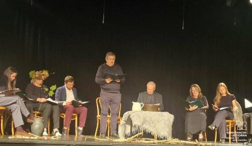 Zdjęcie z czytania Dziadów na ciemnej scenie. Grupa osób siedzi na krzesłach wzdłuż sceny, trzymając w rękach egzemplarze tekstu, podczas gdy mężczyzna na środku czyta na stojąco. Przed nimi na podłodze leżą rozrzucone gałęzie, a pośrodku stoi mały stolik przykryty jasnym obrusem z dzbankiem i misą.