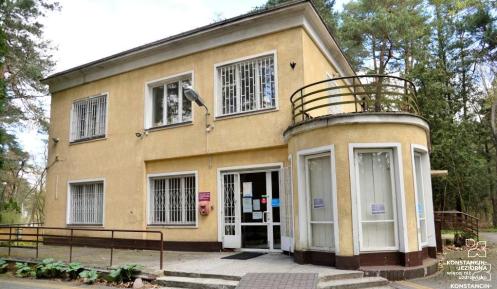 Budynek przychodni wśród drzew, z żółtą elewacją, dużymi oknami z kratami oraz zaokrąglonym wejściem z podjazdem dla osób z niepełnosprawnościami.