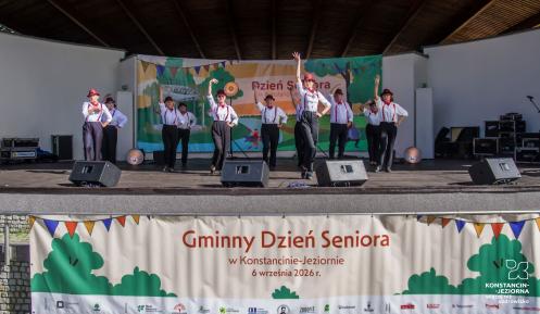 Występ artystyczny seniorów na scenie amfiteatru podczas Gminnego Dnia Seniora w Konstancinie-Jeziornie. Grupa tancerzy w białych koszulach, ciemnych spodniach, czerwonych szelkach i kapeluszach wykonuje układ taneczny z uniesionymi rękami. Z przodu sceny wisi duży baner z napisem „Gminny Dzień Seniora w Konstancinie-Jeziornie”.