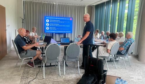 Seniorzy uczestniczą w szkoleniu z cyberbezpieczeństwa. Prelegent wyjaśnia kroki postępowania w przypadku oszustwa, widoczne na slajdzie w tle.