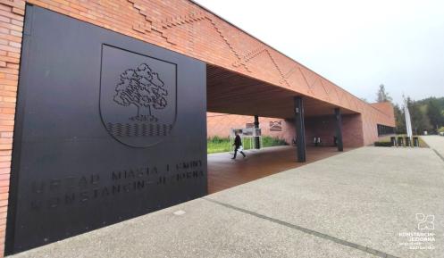 Budynek prostokątny, ceglana czerwona elewacja, dużo okien, płaski dach. Przed budynkiem rosną krzewy, jedno duże drzewo i trawy ozdobne. 