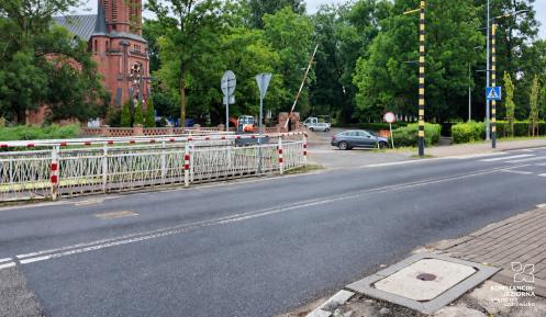 Most na ul. Mirkowskiej z zamkniętym przejazdem kolejowym, na tle zieleni i zabudowy, z widocznym oznakowaniem drogowym i przejściem dla pieszych.