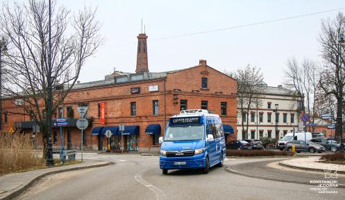Niebieski autobus linii L21 jedzie ulicą na tle zabytkowego, ceglanego budynku w centrum miasta.