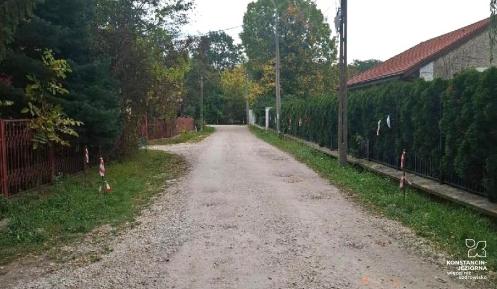 Droga gruntowa z kruszywa, biegnąca między ogrodzonymi posesjami, z drzewami i zielenią po obu stronach oraz widoczną infrastrukturą energetyczną.