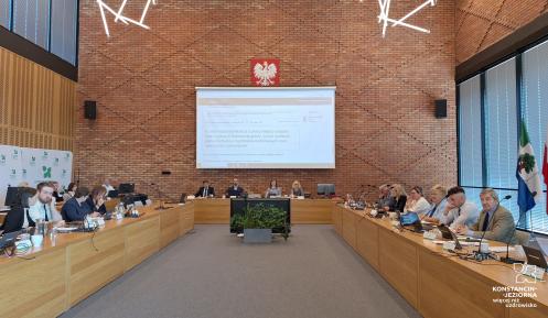 Grupa osób siedzi za stołami na sali posiedzeń