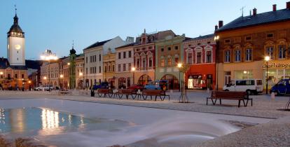 Widok na rynek miejski, z zabytkową zabudową, po prawej stronie szereg kamieniczek, na lewo fragment budowli sakralnej z wysoką wieżą. W centralnej części znajduje się duża fontanna.