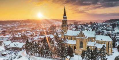 Widok z góry na pokryte śniegiem miasto. W centralnej części, pośród drzew iglastych znajduje się kościół z wysoką wieżą. W tle panorama gór oraz wielobarwne niebo  z zachodzącym słońcem.