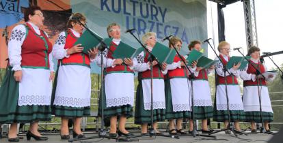Zdjęcie przedstawia zespół Łurzycanki występujący na scenie podczas Jarmarku Kultury Urzecza. Osiem pań, ubranych jest w tradycyjne stroje z regionu. W rękach trzymają śpiewniki. 