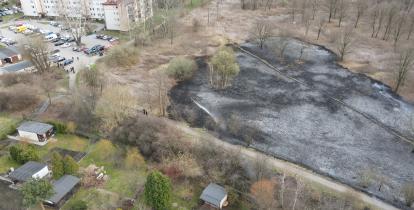 Widok z lotu ptaka, z lewej widoczne bloki mieszkalne, po prawej duży obszar wypalonej trawy, na dole widoczne małe domki na ogródkach działkowych