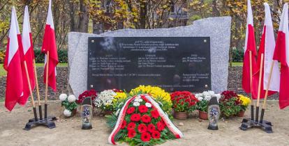 Pomnik kamienny z tablicą marmurową, po prawej i lewej stronie flagi Polskie na drzewcach w stojakach, przed pomnikiem kwiaty i znicze