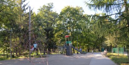  Plac zabaw z dwoma dużymi urządzeniami: do wspinania oraz zjeżdżania. Bawi się na nim kilkoro dzieci. Wokół rosną wysokie drzewa. 