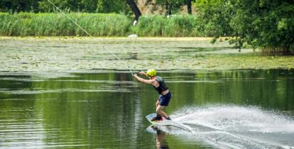 Staw, na środku którego mężczyzna uprawia wakeboarding. Ma na sobie kamizelkę ratunkową, kask na głowie, na nogach przypiętą podłużną deskę, dzięki której może ślizgać się po tafli wody. W rękach trzyma uchwyt połączony z liną, która go ciągnie. 