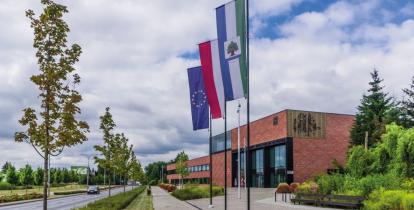 Duży, nowoczesny budynek użyteczności publicznej  ukazany z dużej perspektywy narożnika, ceglane ściany, duże okna i oszklenia, przed budynkiem drzewa oraz horągiew Konstancina-Jeziorny, flagi Polski i Unii Europejskiej na masztach 