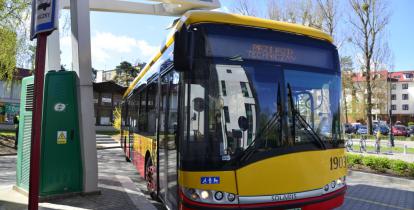 Żółto-czerwony autobus stoi pod wysięgnikiem stacji do ładowania. Z lewej strony znak drogowy. Tło stanowi zabudowa wielorodzinna.