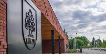 Z lewej strony fragment budynku z czerwonej cegły oraz umieszczony na metalowej części tłoczony zarys herbu gminy. Z prawej strony w oddali drzewa oraz maszty flagowe. Niebo nad budynkiem jest zachmurzone.