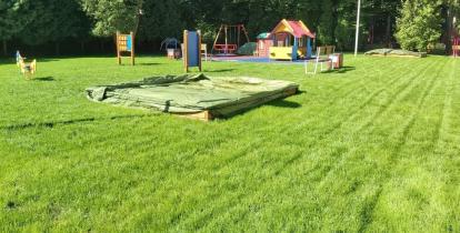 Plac zabaw dla dzieci z zabawkami i urządzeniami, pokryty nowym trawnikiem, pośrodku przykrtya folią piaskownica
