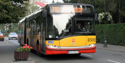 Przód autobusu miejskiego linii 710, jadącego po ulicy, z prawej strony chodnik na którym stoją donice z kwiatami i latarnie