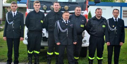 Siedmiu mężczyzn stoi w mundurach strażackich na tle wozu strażackiego.