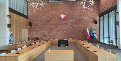 Duża sala. Na środku znajdują się stoły ułożone w kształcie litery U. W głębi widać ceglaną ścianę, na której wisi godło Polski. 