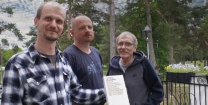 Trzech mężczyzn stoi obok siebie, jeden trzyma w ręku książkę, którą wspólnie napisali. Od lewej: Adam Zyszczyk, Paweł Komosa oraz Witold Rawski. W tle widać wysokie zielone drzewa.