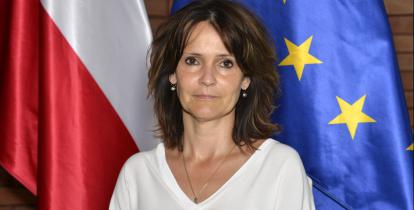 Zdjęcie portretowe kobiety – ciemne proste włosy, ubrana w jasną bluzkę. W tle flaga Polski i Unii Europejskiej.