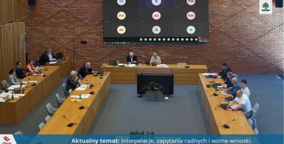 Sala posiedzeń urzędu. Przy dużych drewnianych stołach siedzą osoby obradujące. W tle, na rzutniku wwyświetlone są ikonki osób obradujących zdalnie.