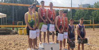 Podium sportowe. Na podestach z numerem 1,2,3 stoją w parach zawodnicy. Na szyi mają medale. W rękach torby z upominkami. W tle widać boisko do siatkówki plażowej. 