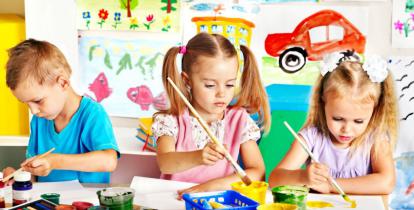 Trójka przedszkolaków siedzi przy stoliku i maluje kolorowymi farbami. W tle galeria z dziecięcymi pracami.