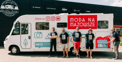 Pięciu mężczyzn, w czarno-białych koszulkach i okularach przeciwsłonecznych, stoi na tle samochodu typu kamper z logo Moda na Mazowsze. Jeden z nich wskazuje na napis: #KamperemPoMazowszu.