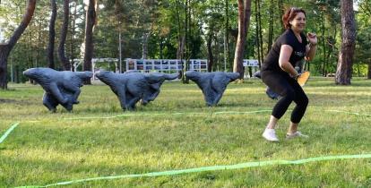 Kobieta w czarnej koszulce i czarnych spodniach podczas gry w crossmintona w parku. W lewej ręce trzyma rakietkę, prawą – ma ugiętą i skierowaną do góry. W tle cztery rzeźby biegnących wilków.