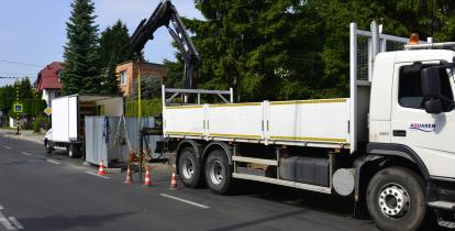 Po prawej stronie samochód ciężarowy z wysięgnikiem, stojący na jednym pasie jezdni. Za nim ustawione są pomarańczowo-białe pachołki drogowe odgradzające właz studzienki kanalizacyjnej. Na tym samym pasie stoi samochód dostawczy z otwartymi tylnymi drzwiami. W tle drzewa i osiedle domków jednorodzinnych.