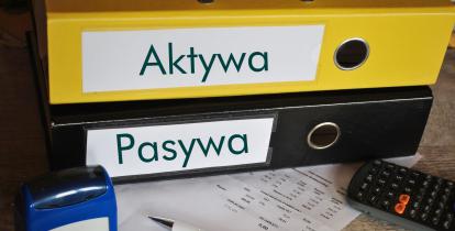 Na biurku leżą dwa segregatory podpisane: aktywa, pasywa.  Obok nich znajduje się pieczątka, długopis oraz kalkulator. 