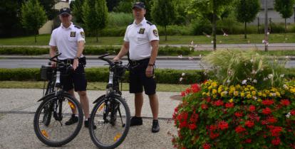 Dwaj mężczyźni, strażnicy miejscy, w służbowych strojach stoją przy rowerach. W tle drzewa i droga. Po prawej stronie wieża z kwiatami.