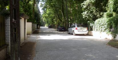 Widok na ulicę o nawierzchni z kostki brukowej, w oddali na poboczu stoją dwa samochody osobowe. Po prawej i lewej stronie znajdują się ogrodzenia posesji. 
